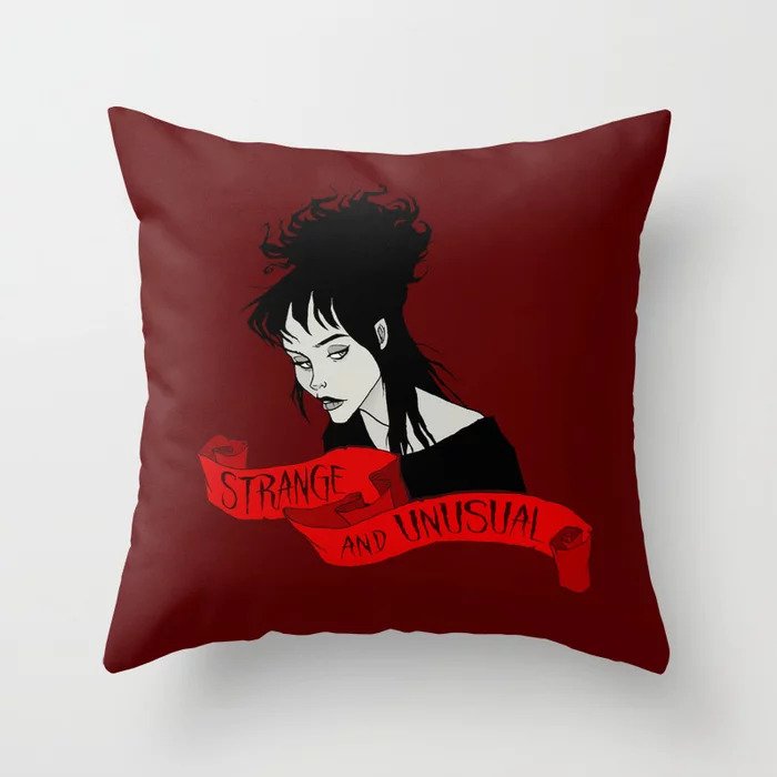 Photo of burgundy throw pillow with drawing of Lydia Deetz with a ribbon underneath with the words strange and unusual on it.